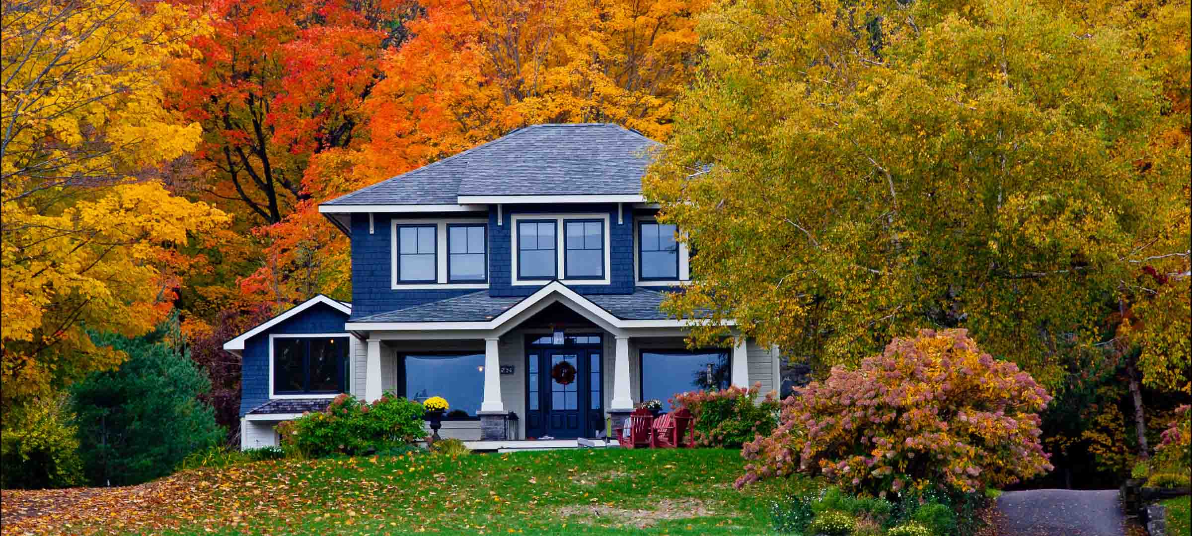 Blue-and-white two-story house surrounded by vibrant red, orange, and yellow autumn trees. Blue-and-white two-story house surrounded by vibrant red, orange, and yellow autumn trees.