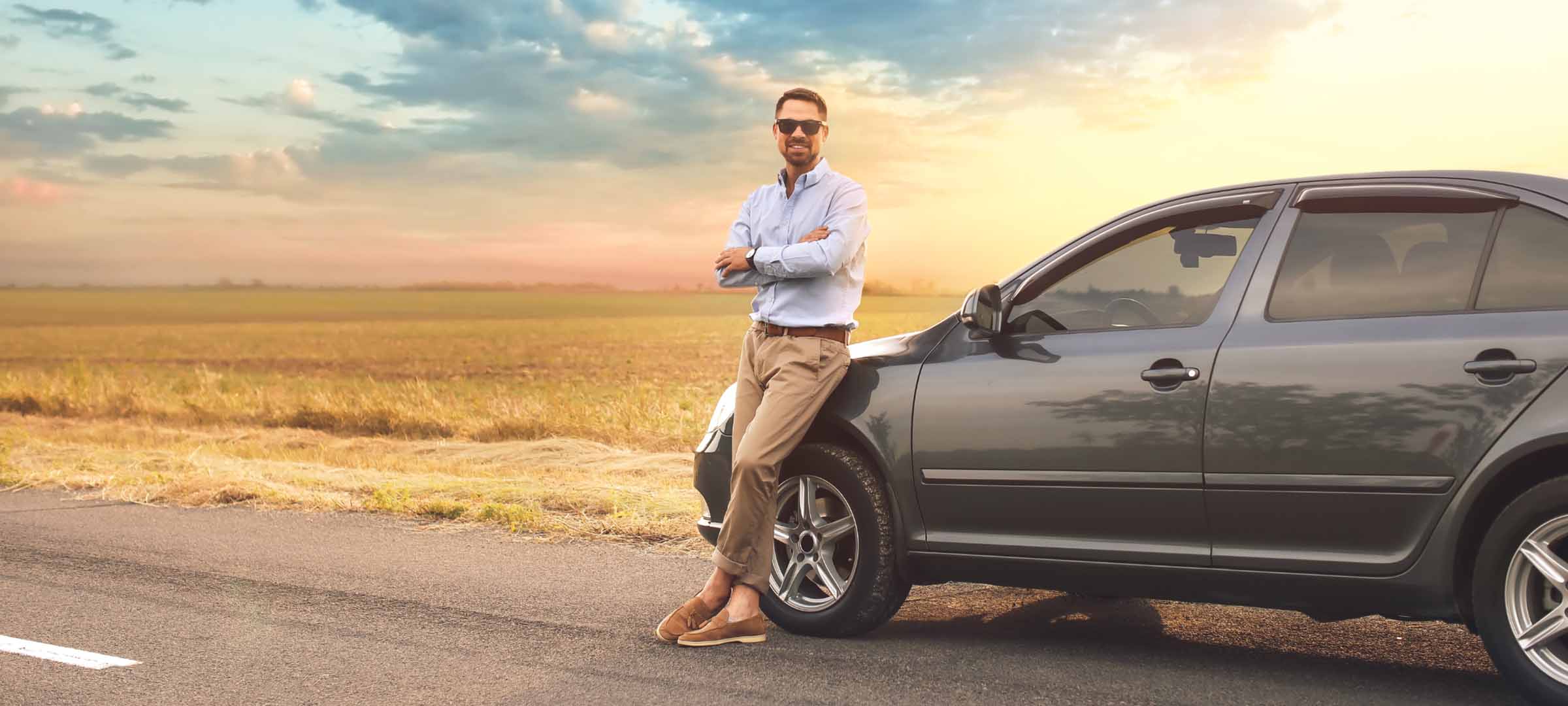 Man leaning against a dark gray sedan on a country road at sunset - https://jamesaryaninsagencyllc.com/