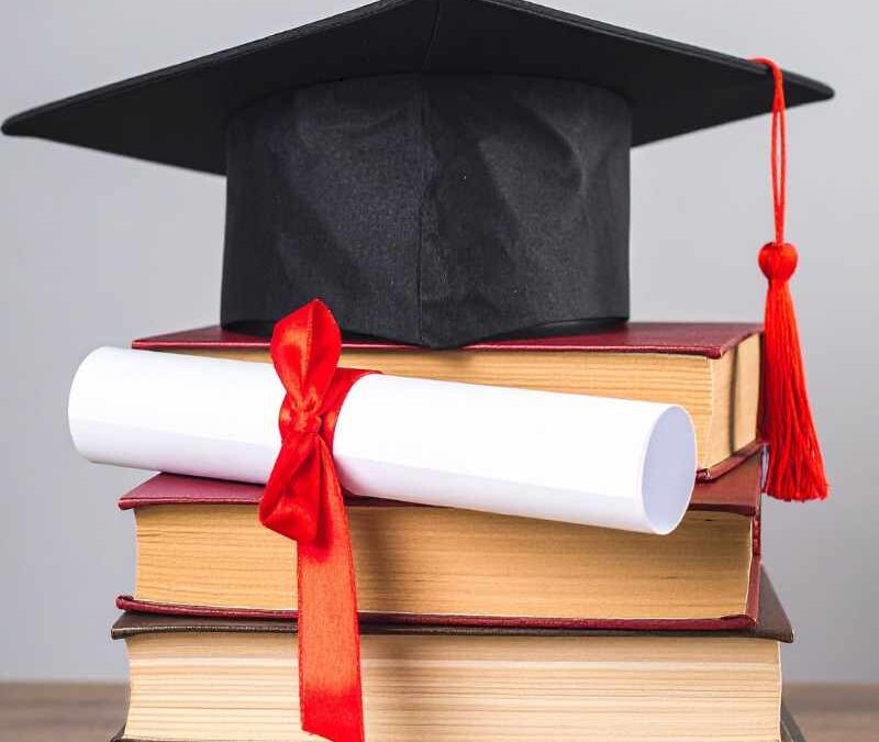 Graduation cap and rolled diploma with red ribbon sit atop a stack of books on a wooden table, symbolizing education and achievement