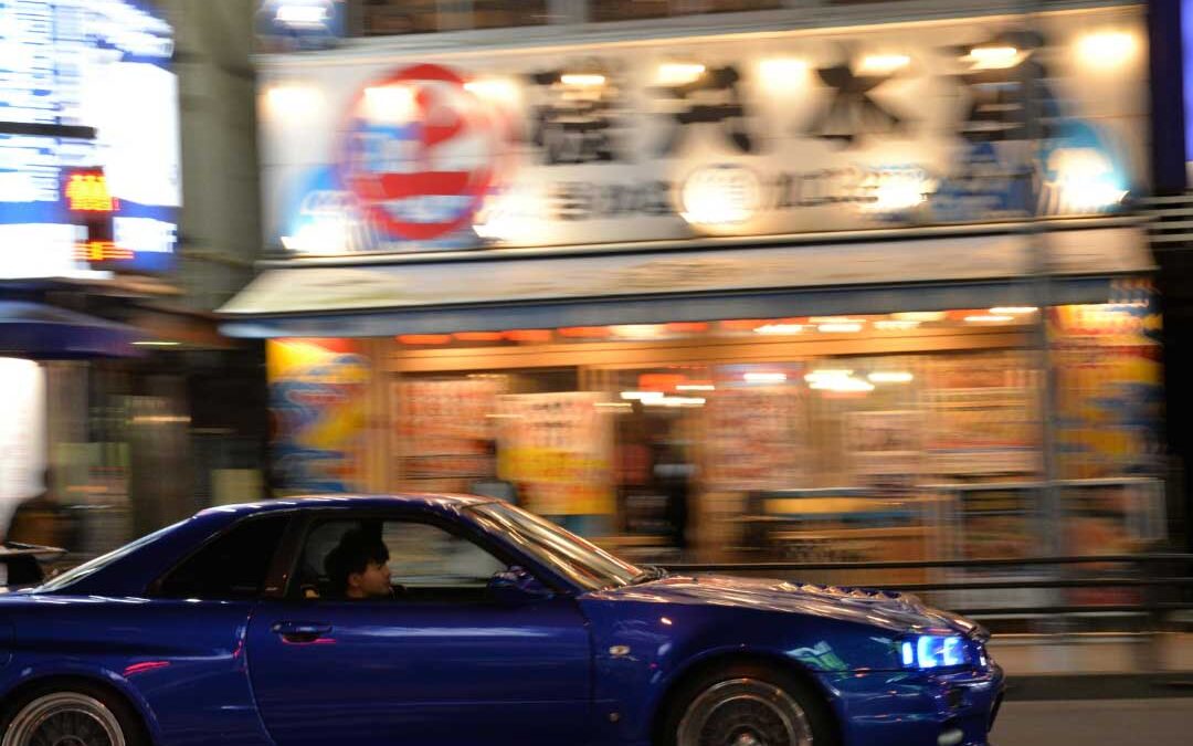 A blue car drives down a dimly lit street at night, illuminated by streetlights