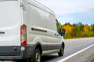 White cargo van driving on a paved road beside autumn trees. White cargo van driving on a paved road beside autumn trees.