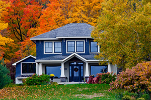 Two-story blue-gray house surrounded by vibrant autumn trees and fallen leaves.