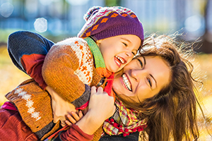 A smiling child hugs their mother outdoors on a sunny day.